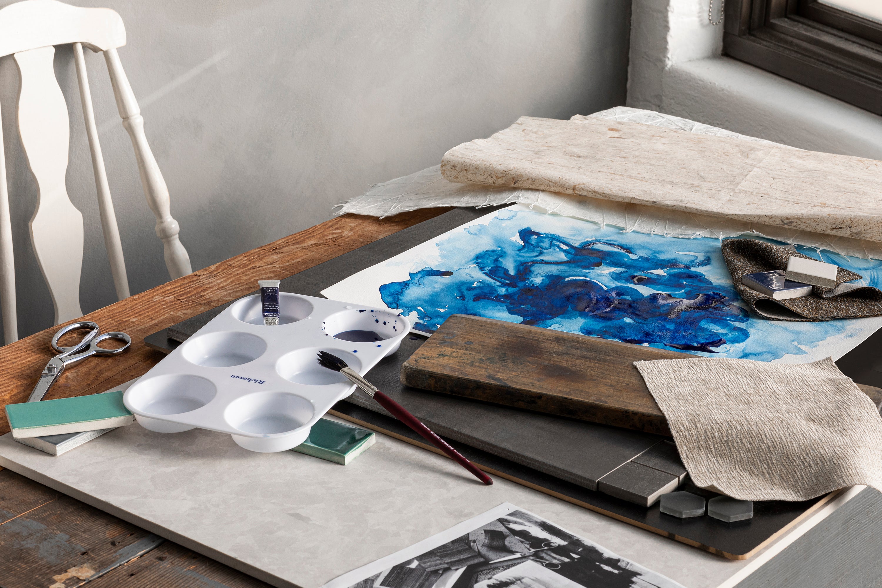 Wooden table covered with watercolor supplies and watercolor painting in blue.